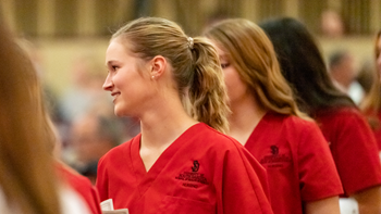 Three nursing students walk in a line. The front student looks to their right. The students are wearing red scrub shirts.