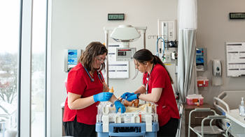 Two nursing students observe and work on a medical dummy. The students are wearing red scrubs and stand facing each other over an infant bed in a simulation lab.