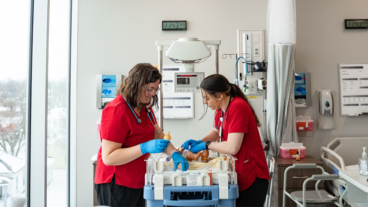 Two nursing students observe and work on a medical dummy. The students are wearing red scrubs and stand facing each other over an infant bed in a simulation lab. 