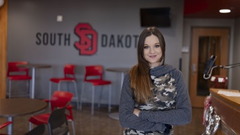 USD - Sioux Falls pre-nursing student Sadi Peck stands in front of a USD sign with her arms crossed. She is smiling at the camera.