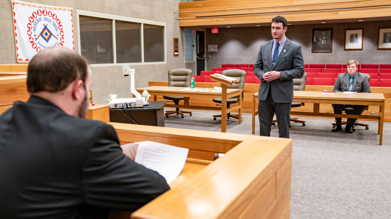 A student stands facing forward and presents to another student sitting in a judge stand. They are in a courtroom. Another student sits in the background and observes.