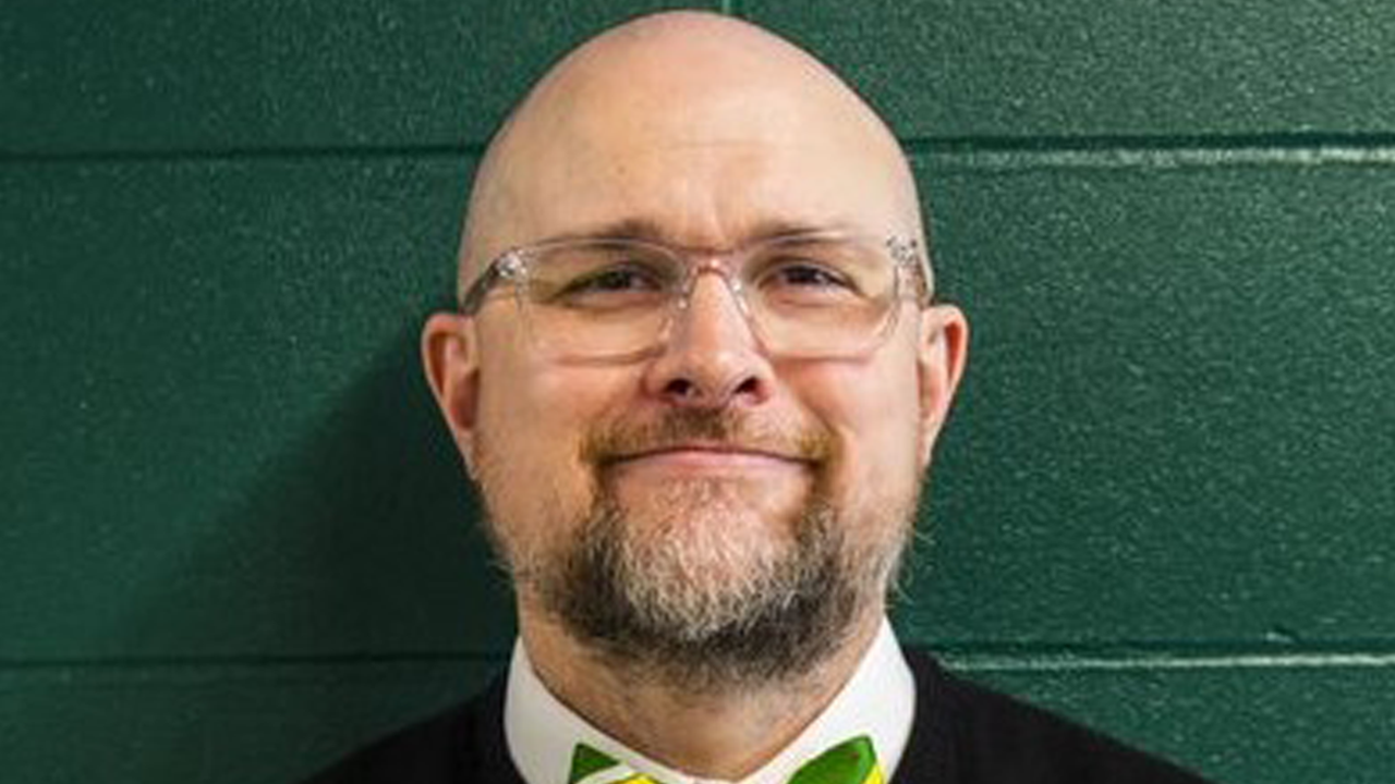 George Hawkins stands against a dark green wall and smiles for a photo.