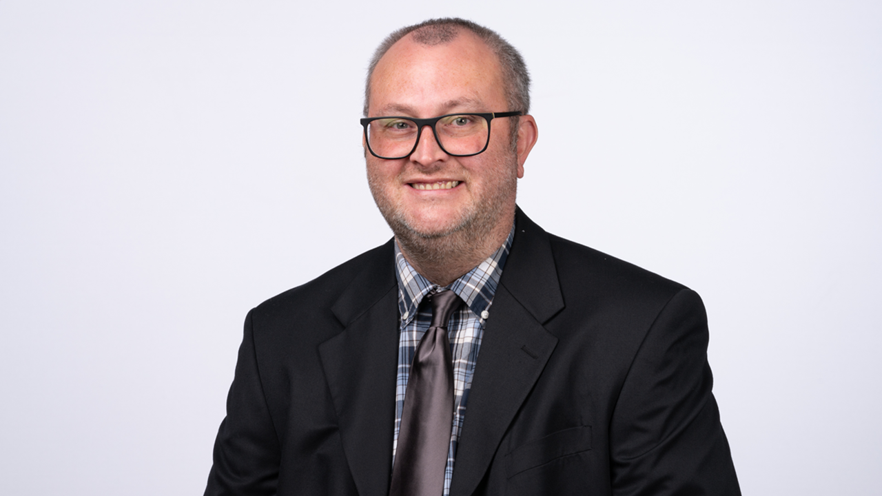 A headshot of Justin Huston. He is wearing a suit and glasses and is sitting in front of a white background.