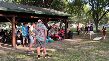 College students mingle at a picnic in the park. A majority of the students sit under a roof and enjoy food at picnic tables. A few students stand in the grass and play cornhole.