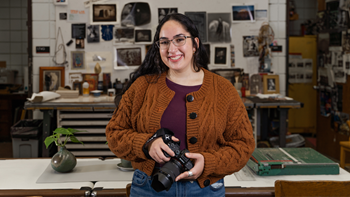 A female student stands in an art studio and holds a camera in her hands.