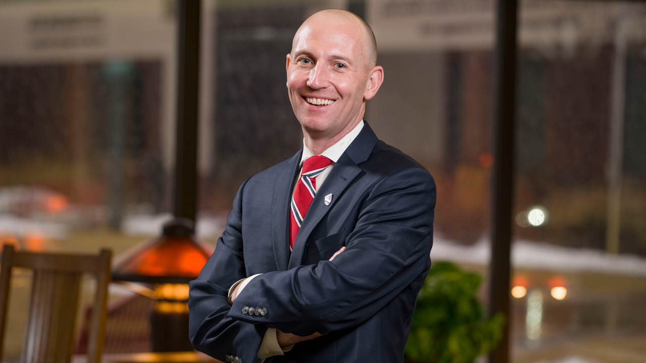 Neil Fulton wears a suit and tie and smiles with his arms crossed. He stands in the law school, with a dim lamp lit behind him.