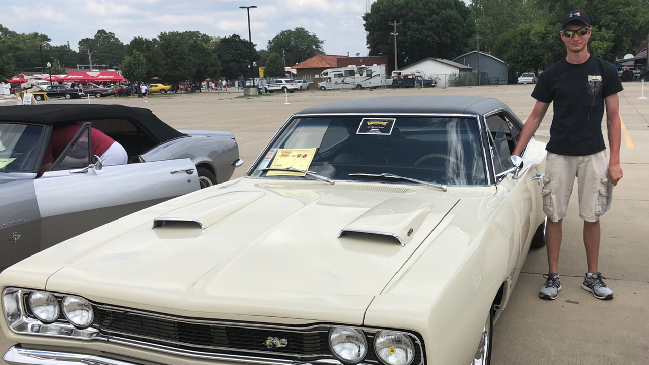 A persons stands next to a tan-colored classic car.