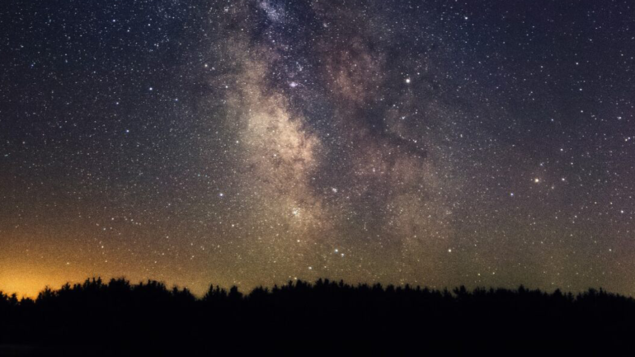 A night sky with a line of trees at the bottom and glowing purple, orange and yellow colors and stars in the sky.