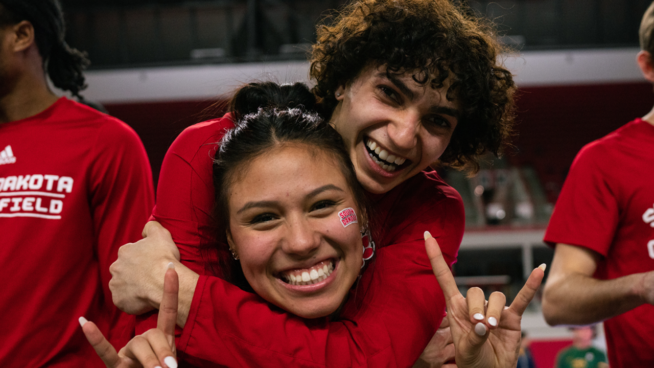 Two student-athletes wearing red smile for a picture. One athlete stands behind the other and hugs them for the picture.
