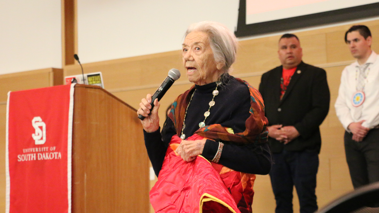 Elizabeth Cook-Lynn holds a microphone and talks near a podium. In her other hand, she holds a Star Quilt.