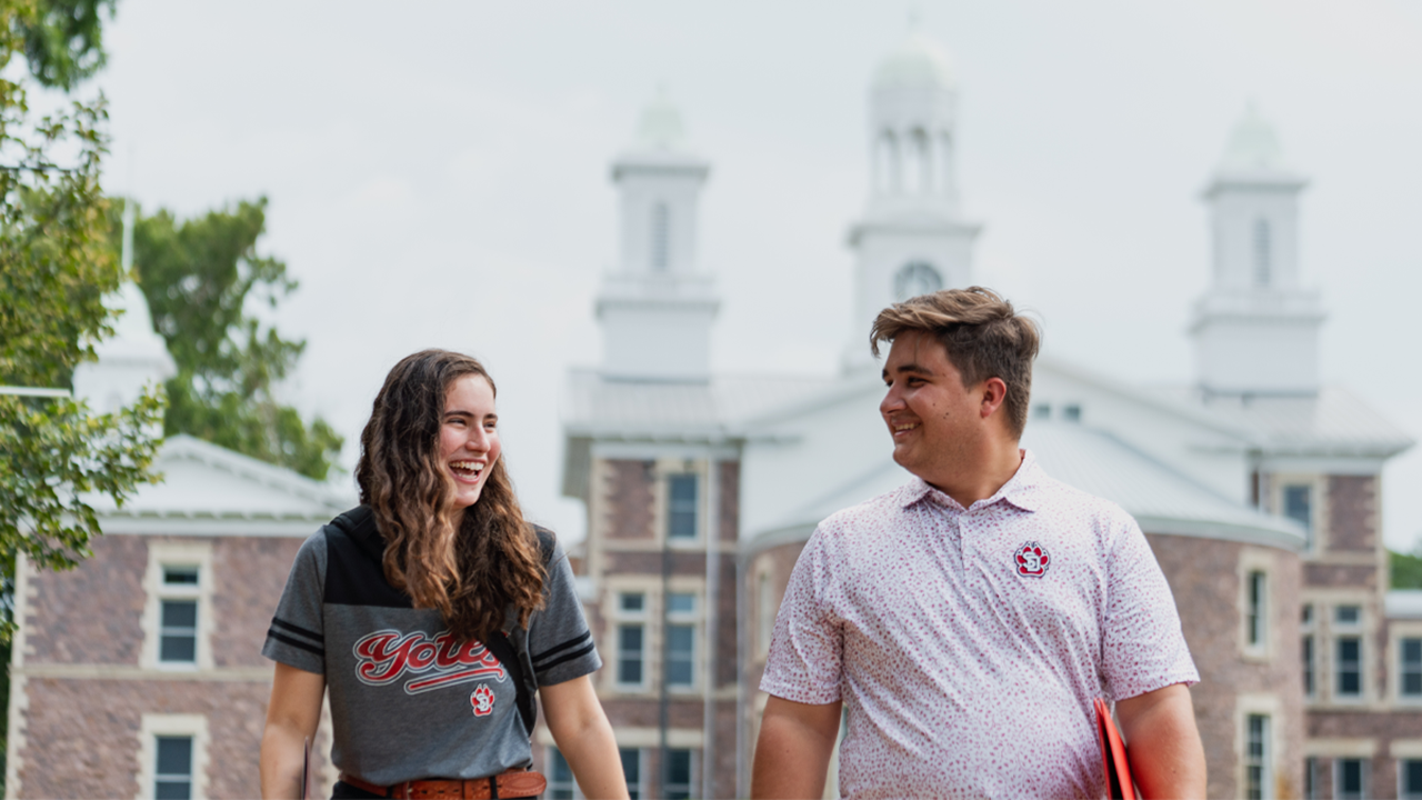 Two students walk and talk while smiling at each other. Behind them is USD's iconic Old Main building.