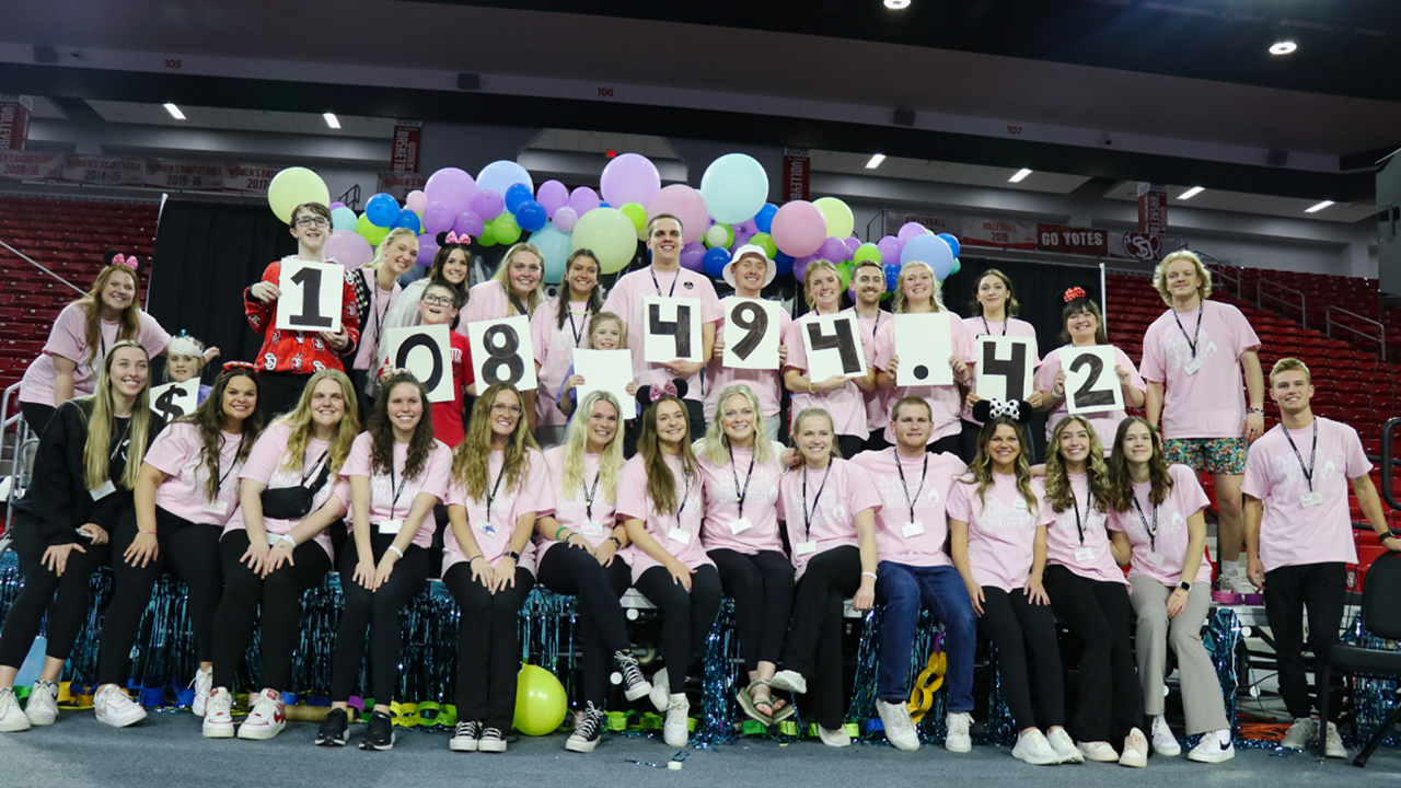 A group of over 20 college students stand together and hold up pieces of paper with the number 108,494.42. There are balloons behind them and some children standing amongst them. They are all wearing pink shirts.