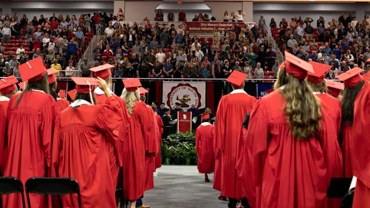 Students in red graduation gowns and caps stand and face the podium and crowd at a commencement ceremony.