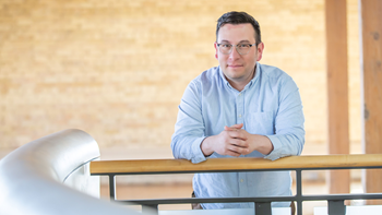 Joe Williams leans against a railing in front of a light colored brick background.