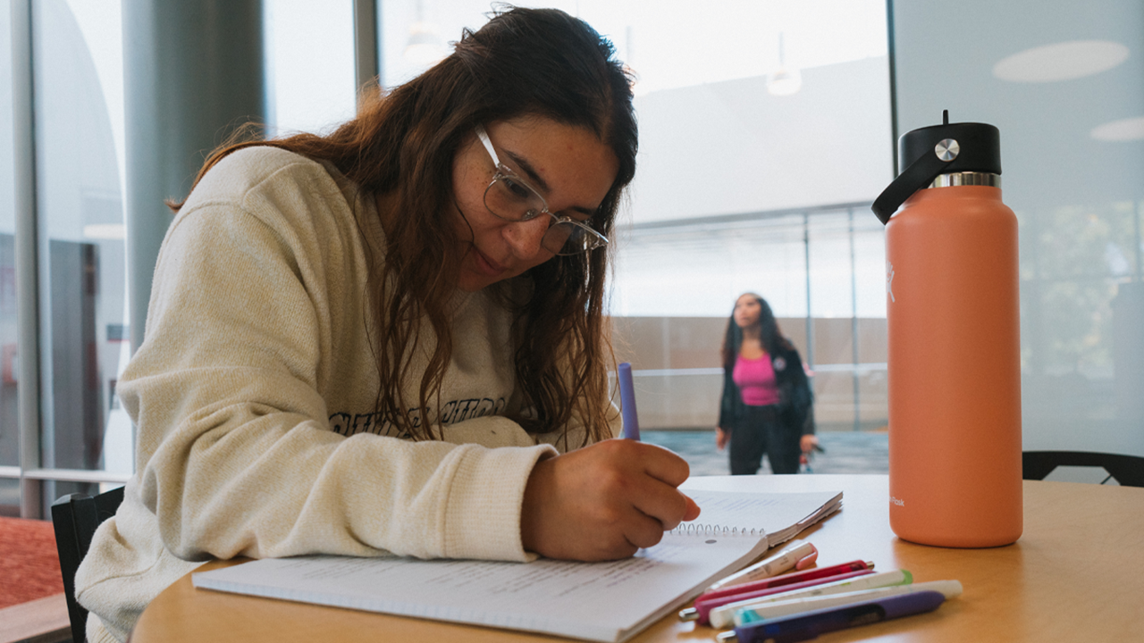 A student looks down at a piece of paper and writes on it. Another student walks in the background.