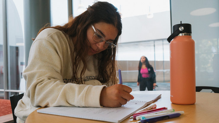 A student looks down at a piece of paper and writes on it. Another student walks in the background. 