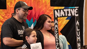 Three people pose in front of a quilt with a frame that says "Native American Alumni."
