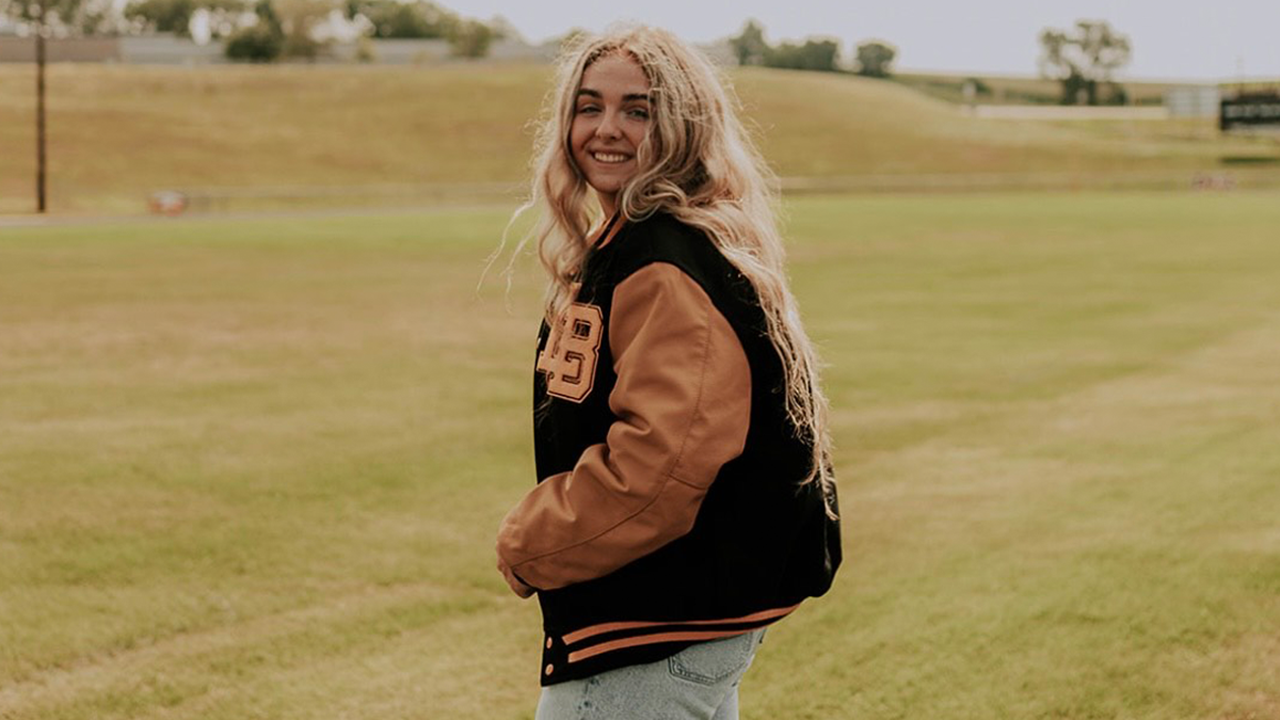 A female student wearing a letterman's jacket and standing in a field of grass. Her body faces toward the left of the photo with her face looking at the camera.