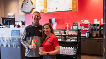 Travis and Kristen Rhoades stand with their arms around each other and coffees in their hands. They stand in front of a cashier counter at Scooter's Coffee.