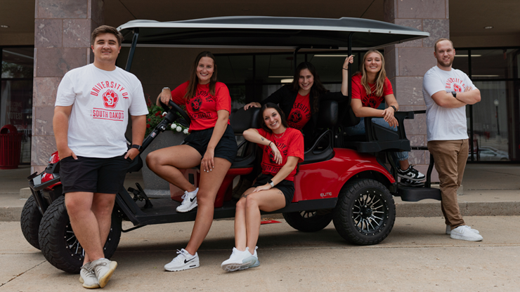 Six student ambassadors sit on a Charlie Cart in front of the Wagner Welcome Center.