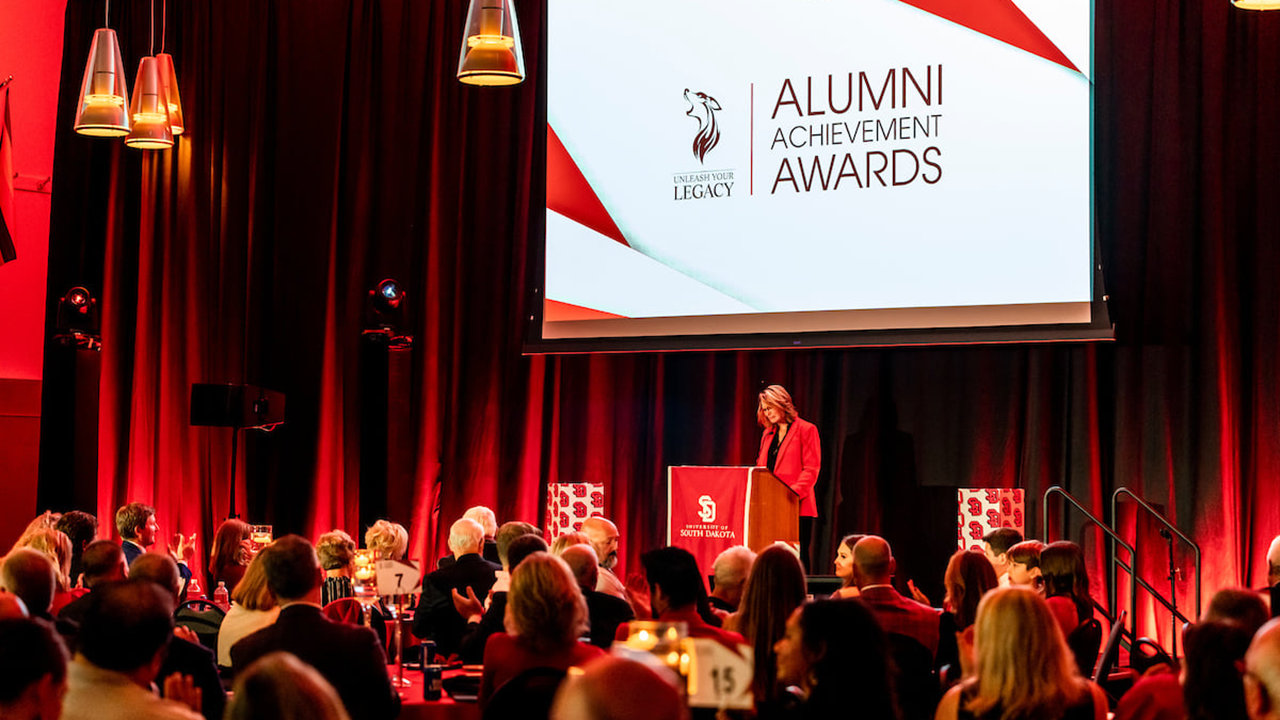 A darkly lit room with red lighting accents is filled with people sitting around tables. A person in a red blazer stands at a podium and speaks to the room.