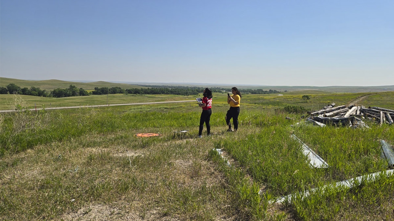 Two students are in a South Dakota field, gazing toward the sky at their drone taking footage of the field. Two students are in a South Dakota field, gazing toward the sky at their drone taking footage of the field.