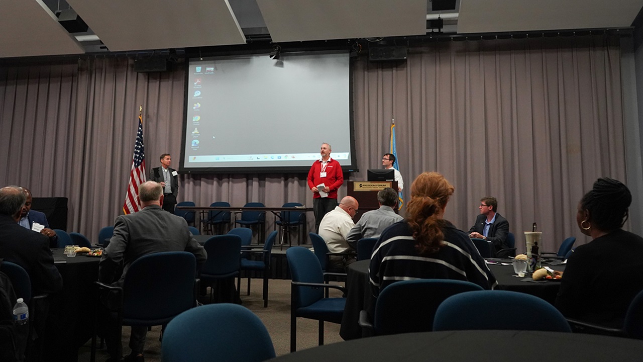 A view from the audience of a stage with people presenting on a projector screen.