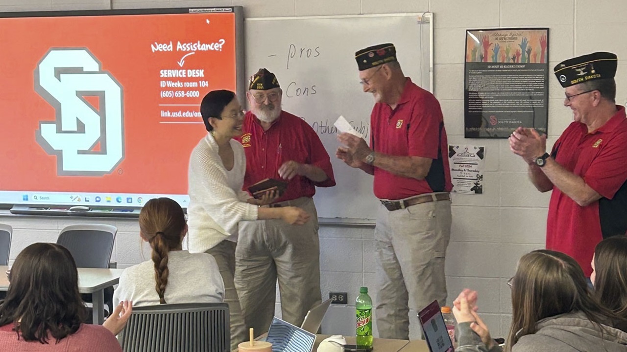 Jing Williams receives a plaque from VFW Post 3061 members in a classroom. They are smiling and shaking hands. Jing Williams receives a plaque from VFW Post 3061 members in a classroom. They are smiling and shaking hands.