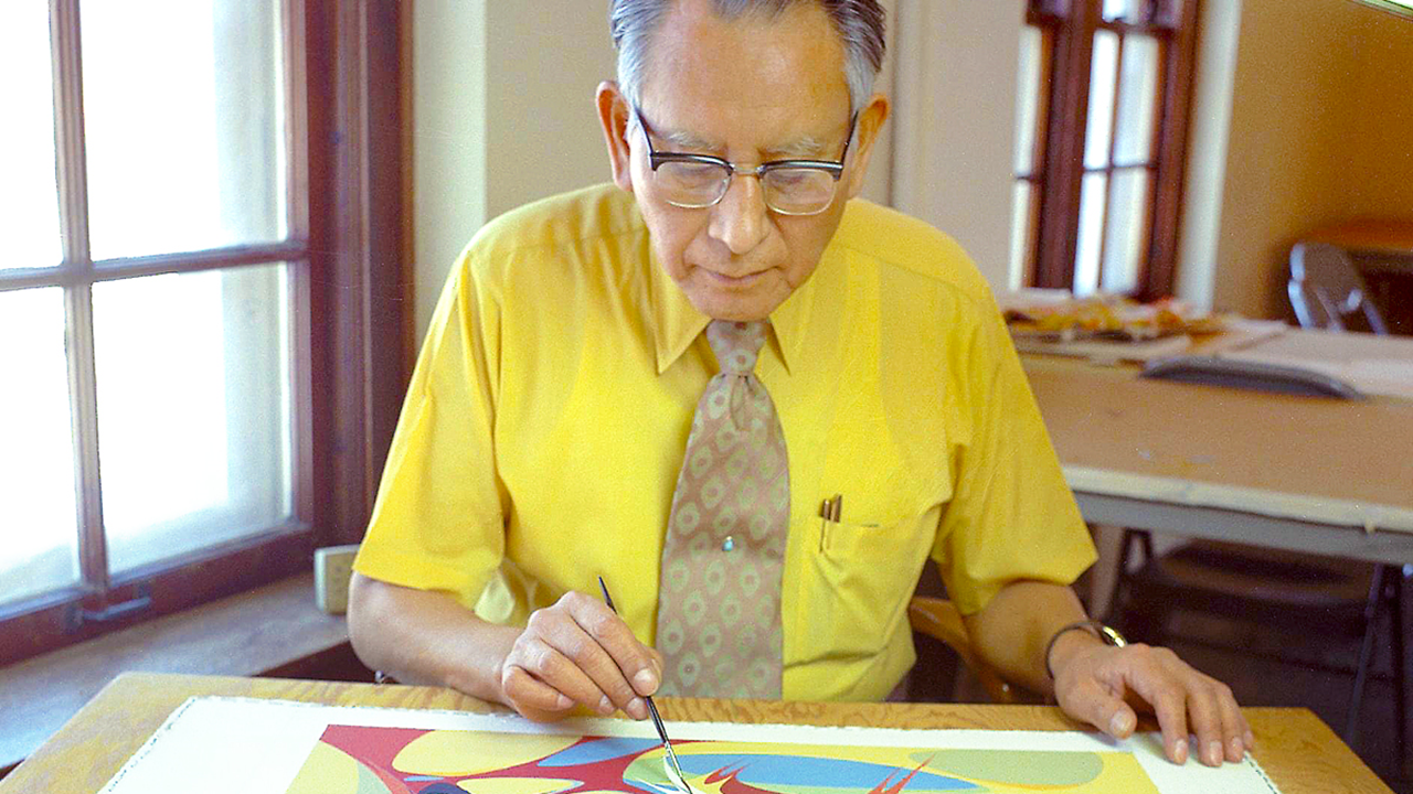 Oscar Howe sits at a table painting a piece of artwork.