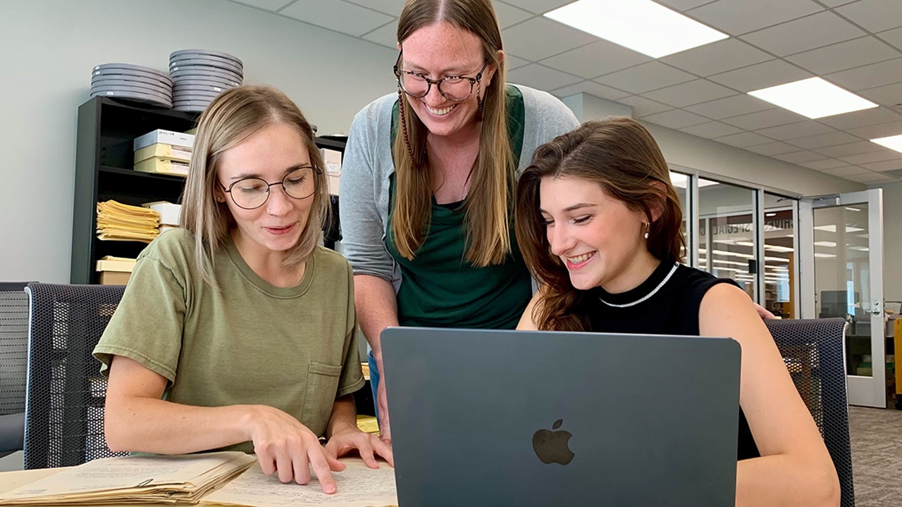 Three women at a table look at a historical document while working on a laptop.