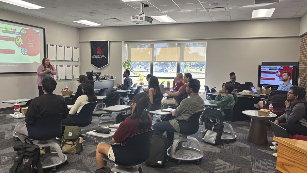 A group of graduate students sitting in desks look to the front of the classroom where a presenter is giving their talk. A group of graduate students sitting in desks look to the front of the classroom where a presenter is giving their talk.