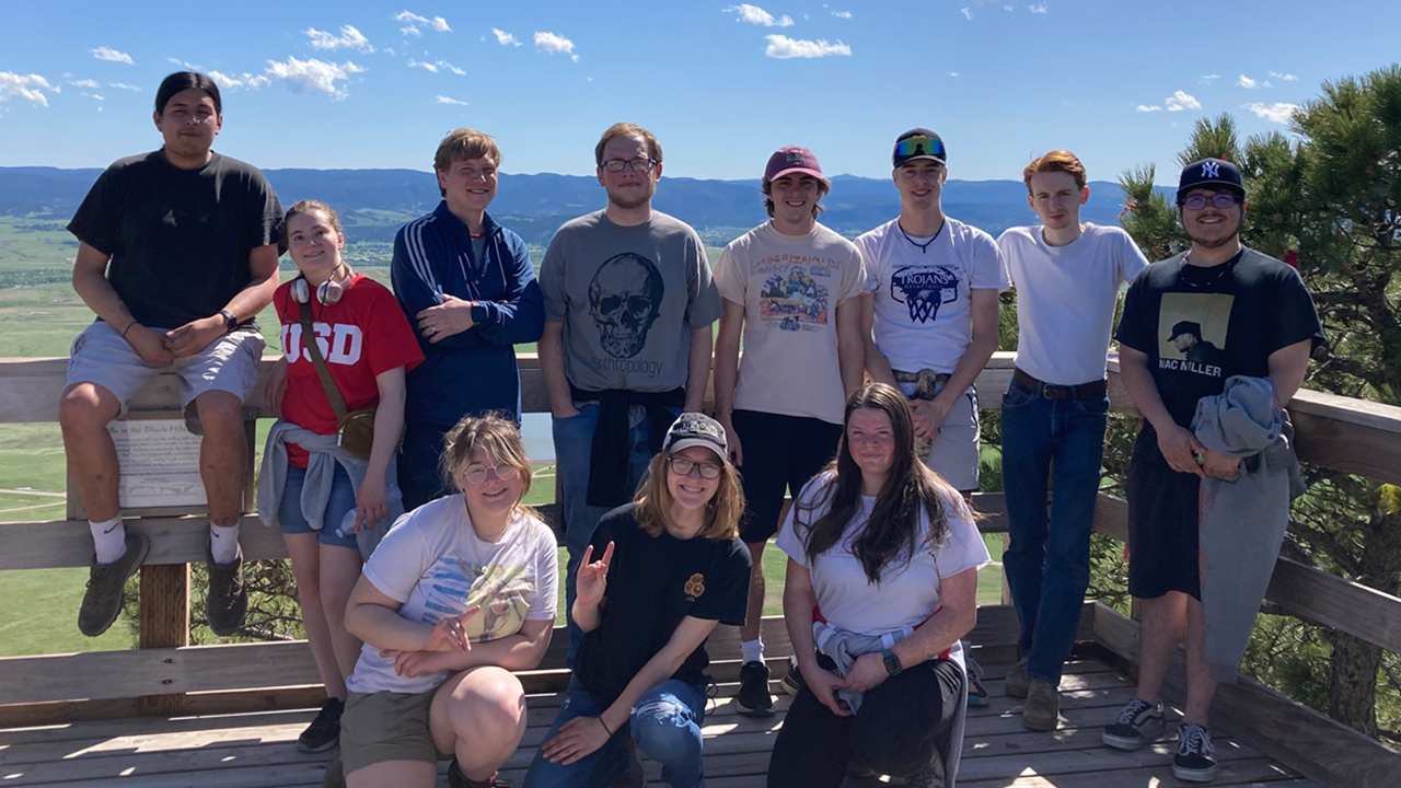 Students and Tony Krus stand together for a photo on a wooden deck overlooking green hills and blue skies.