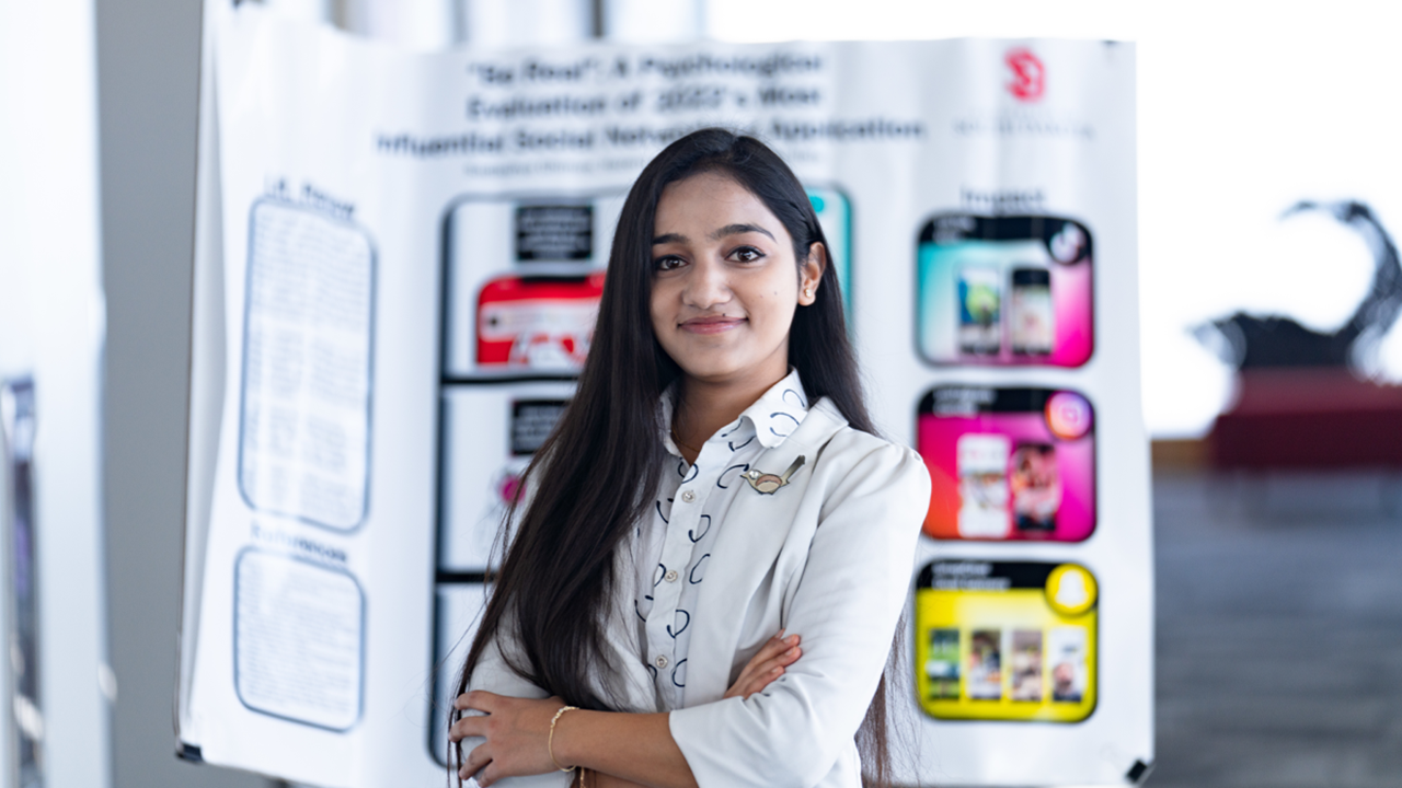 Sadikshya Ghimire stands with her arms crossed in front of her research poster.