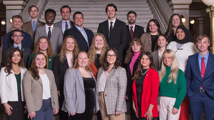 Twenty three college-aged students dressed in professional attire stand together for a group photo on a flight of stairs inside the South Dakota State Capitol.