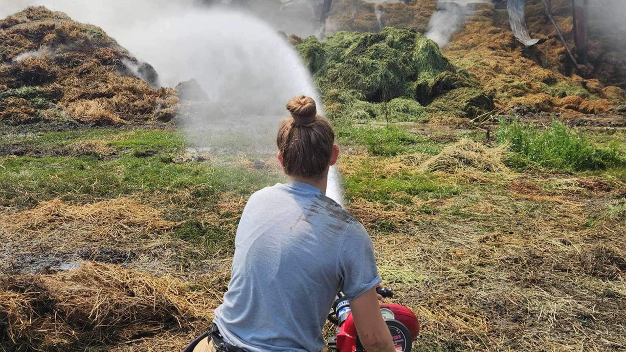 Lilly McNaughton wears a firefighter uniform and sprays a hose onto a big pile of rubble. She is facing away from the camera.
