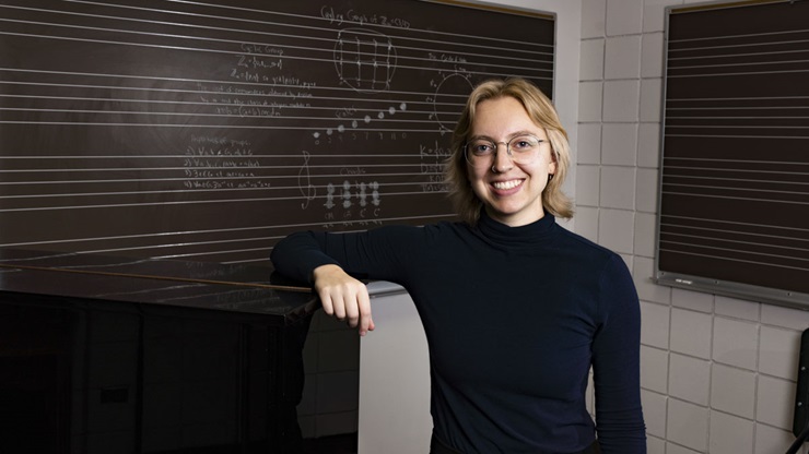 USD student Olivia Roberts leans on a grand piano. She stands in front of a chalkboard with mathematical equations and music notes.
