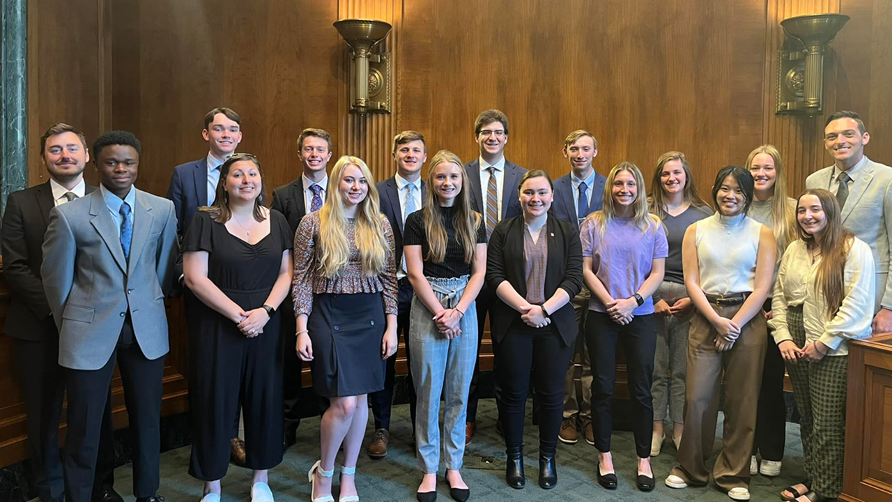 A group of 17 people stand together for a photo. Some of them are college students, one is a professor and some are USD alumni.