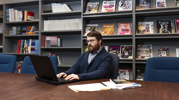 USD professor Travis Loof works on a laptop in an office. Behind him is a shelf of magazines in multiple colors