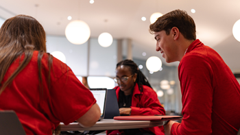 Three business students sit at a table and converse with their laptops opened in front of them.