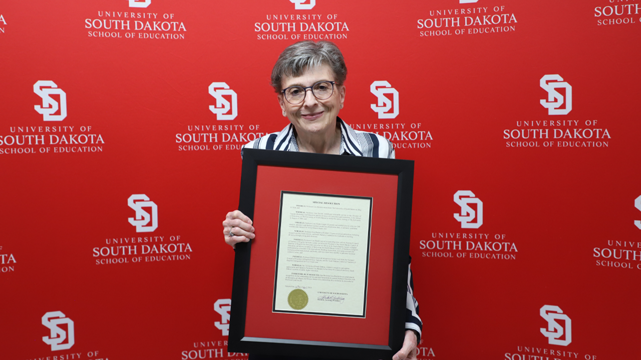 Liza Hazlett smiles and holds her framed emeritus proclamation in front of a red USD School of Education backdrop.
