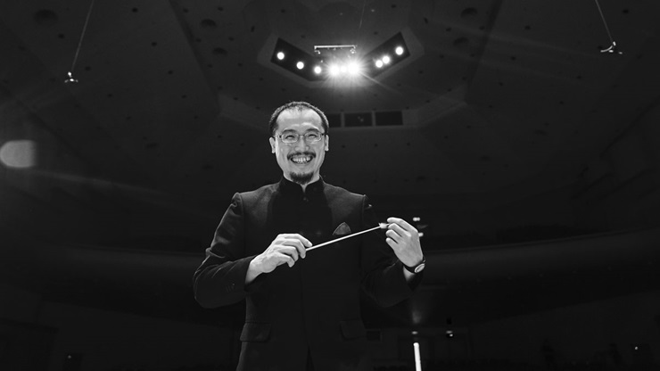 Chun-Ming Chen stands on stage, holding a conductor's baton and smiling at the camera.