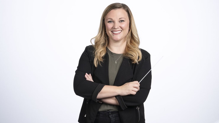 Nicole Decker stands with arms crossed, a conducting baton in one hand. She is smiling. Nicole Decker stands with arms crossed, a conducting baton in one hand. She is smiling.