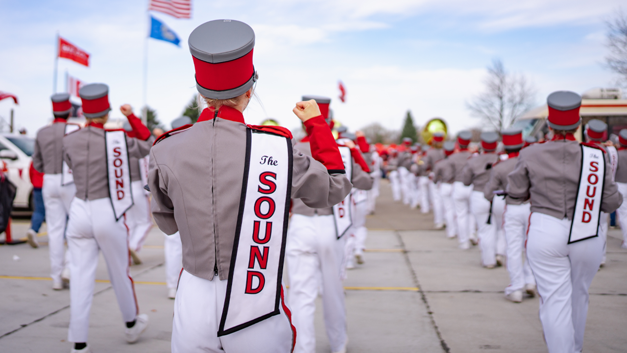 Students in marching band uniforms march forward in formation. They wear sashes that say "The SOUND" on their backs.