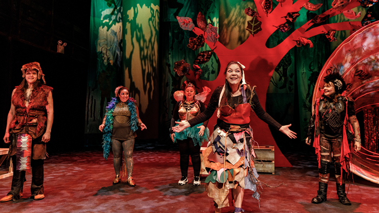 Five actors wear patchwork styled costumes and perform on a stage with a large red tree in the background.