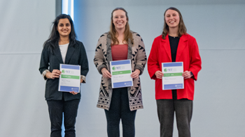 Priyam Pandey, Jennifer Mussell and Morgan Rothschadl stand on stage and hold their first, second and third place 3MT certificates.
