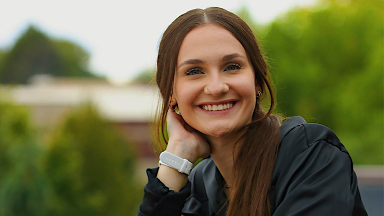 Elle Hoops rests her face in her hand and smiles for a photo. The background is full of vibrant green trees.