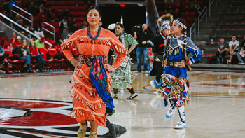 Dancers in colorful regalia perform at halftime of a basketball game on the court.