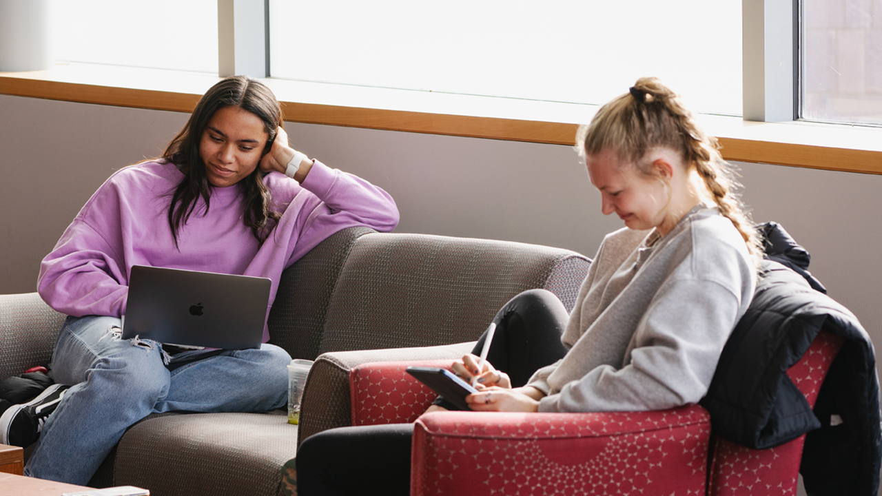 Two students sit in comfy chairs and work on homework.