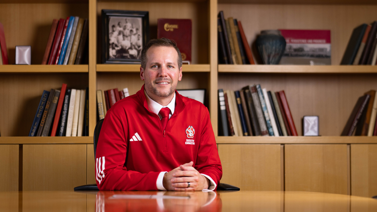 Jon Schemmel sits at a wooden desk with his right arm leaned on the desk. There is a bookshelf behind him.