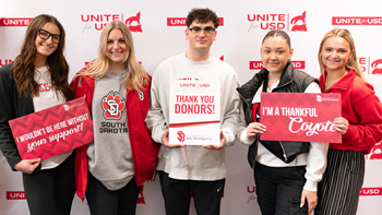 Five students stand together for a photo in front of a white background that says Unite for USD in red text. A few of them hold signs that have phrases like "Thank You Donors" and "I'm a thankful Coyote."